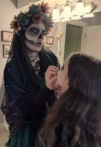 the artist in a dark veil decorated with flowers and her face and neck painted as a skeleton, painting her daughter's face for Halloween