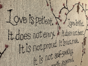 Hand stitched tapestry with tree branches and part of a Bible verse: "Love is patient. Love is kind. It does not envy. It does not boast. It is not proud. It is not rude. It is not self-seeking..."