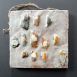 a collection of broken clam and oyster shells of varying gray and tan hues arranged with one smaller shard fastened to a contrasting larger shard, placed in a visually pleasing arrangement on a rough-cut square wood panel with encaustic medium layered over top and visually suggesting wet snow on a beach; a twisted piece of twine is attached with mismatched screws to the top and side to serve as the hanging hardware for this piece