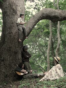 four children dressed up in early 1900s fashion gathered in the woods, one is in a tree and one is strumming a guitar under the tree while the others listen
