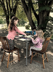 three children sitting at a table outside working on art projects under two shade trees